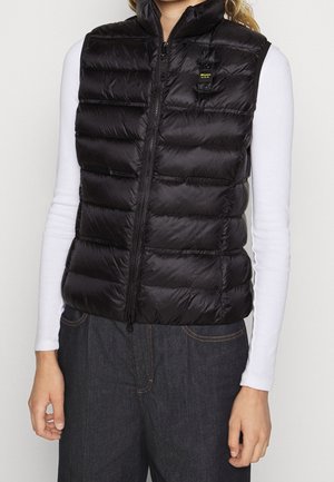 Personne portant un gilet sans manches matelassé noir ajusté sur une chemise blanche à manches longues et un jean foncé, debout les bras détendus.