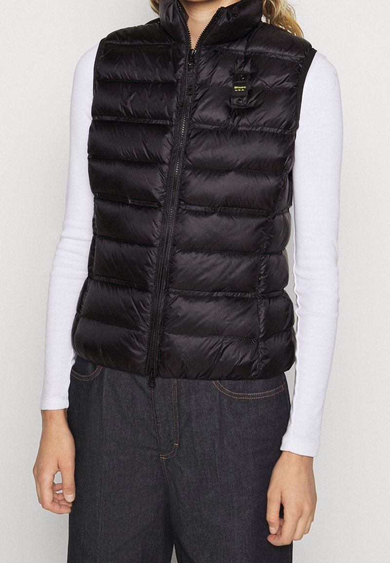 Personne portant un gilet sans manches matelassé noir ajusté sur une chemise blanche à manches longues et un jean foncé, debout les bras détendus.