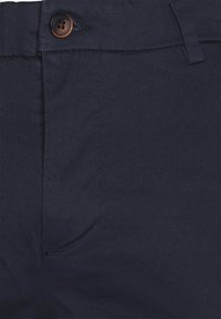 Gros plan sur un pantalon bleu marine foncé montrant le bouton, la ceinture, le rabat de la fermeture éclair, la boucle de ceinture et les détails de couture de la poche avant.