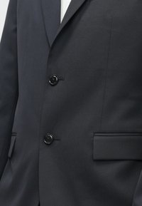 Casaco de fato azul marinho escuro, com corte ajustado, apresentando dois botões pretos e uma textura suave. Tem lapelas cortadas e um único bolso no peito.