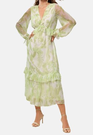 Nainen pukeutuneena läpikuultavaan, pitkähihaiseen, nilkkaan ulottuvaan mekkoon, jossa on vaaleanvihreä kukkakuvio ja röyhelöyksityiskohtia, yhdistettynä beigeihin korkokenkiin.