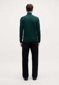 Dunkelgrüner, taillierter Pullover mit einem gerippten Stehkragen, langen Ärmeln und einer glatten Strickstruktur, kombiniert mit schwarzen geraden Hosen.