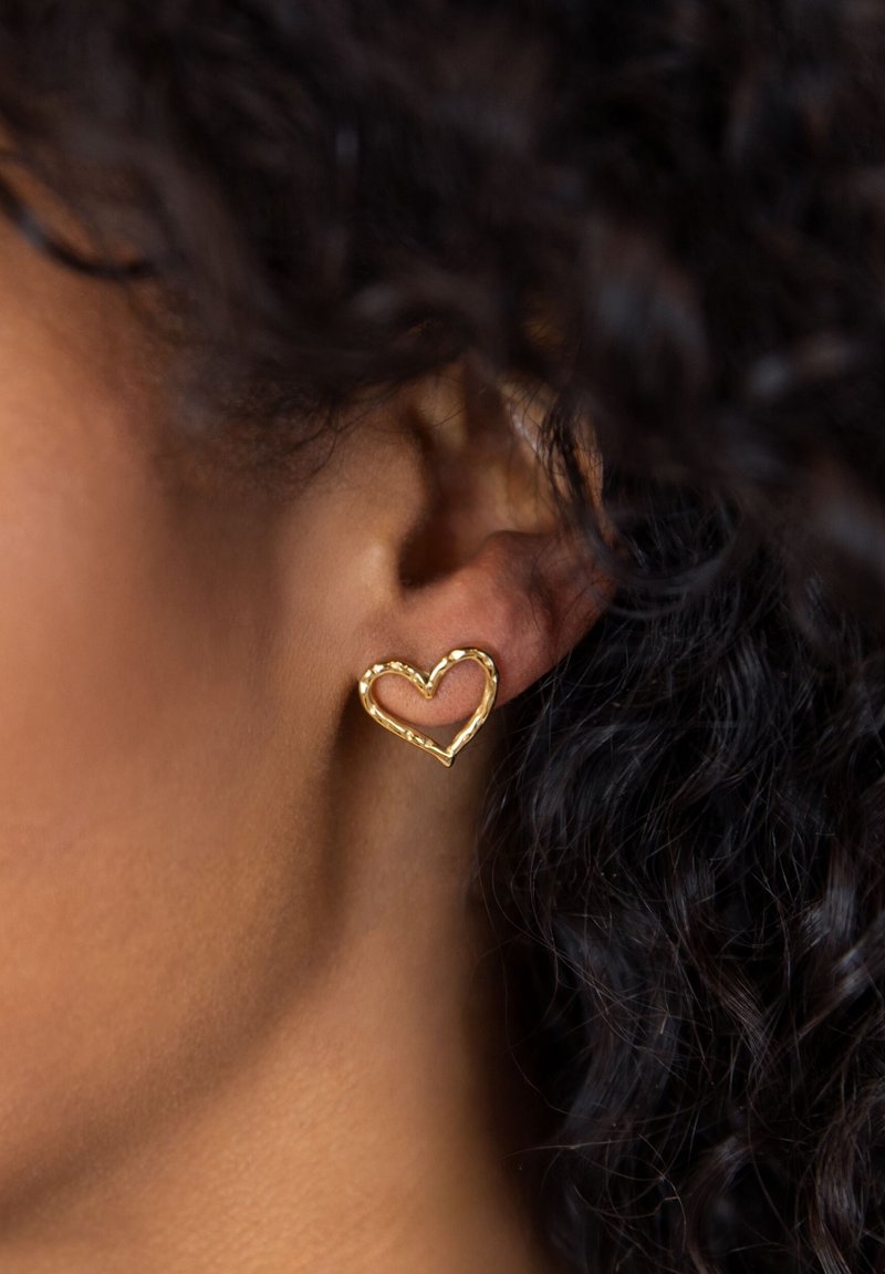 Gouden hartvormige oorbellen met een gestructureerd, open ontwerp, gedragen door een persoon met donker haar en krullen. De oorbellen hebben een minimalistische stijl.