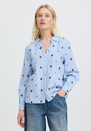 Blonde vrouw draagt een lichtblauw overhemd met zwarte geometrische prints en een blauwe spijkerbroek, staand met één hand in haar zak.