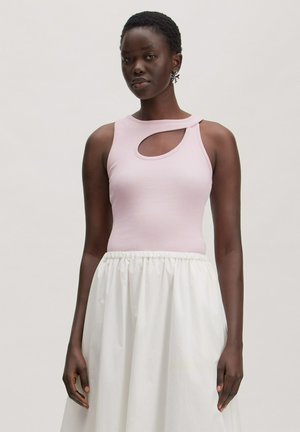 Woman wearing a light pink sleeveless top with a cutout near the neckline and a white gathered skirt against a plain background.