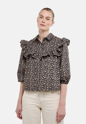 Femme portant un chemisier noir à motifs floraux avec des volants aux épaules et à la poitrine, associé à un pantalon taille haute beige clair, debout devant un fond blanc.