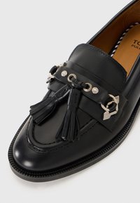 Mocassin en cuir noir avec des détails de pampilles, des œillets argentés et des anneaux en métal décoratifs sur une semelle texturée.