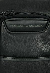 Pochette en cuir noir avec fermeture éclair, surface texturée et logo "Porsche Design" en relief bien visible. Accents horizontaux subtils.