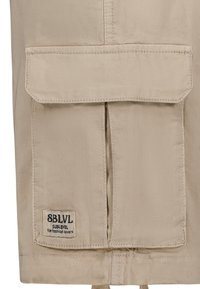 Beige stof met een voorflapzak en een label. De textuur is glad, met topsstikdetails en een koordje aan de onderrand.