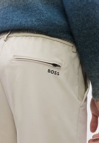 Beiget housut, joissa on sileä pinta, takaosassa vetoketjullinen tasku ja mustalla painettu "BOSS"-logo. Villapaita päällä.