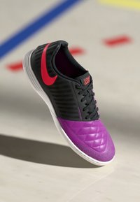 Nike futbola apavi ar melnu augšdaļu, kas rotāta ar violetiem akcentiem, teksturētām rakstu formām, mīkstinātu purngalu un sarkanu logotipu. Balta zole.