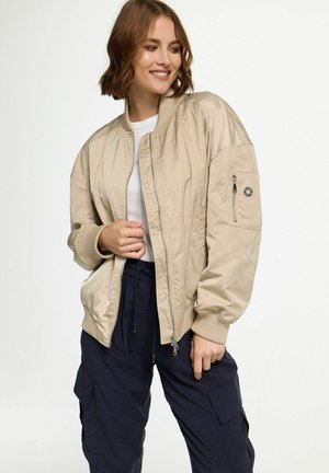 Beige bomberjack gemaakt van soepel, lichtgewicht materiaal met ribbels bij de mouwen, een ritssluiting aan de voorkant en een zijzak met hardwaredetails.