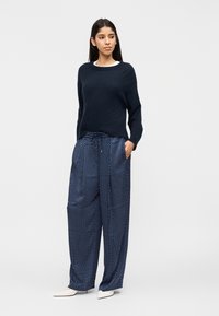 Γυναίκα όρθια με τα χέρια στις τσέπες, φορώντας μπλε σκούρο πουλόβερ, μπλε σκούρα παντελόνια με πουά και άσπρα μυτερά παπούτσια, μπροστά σε απλό φόντο.