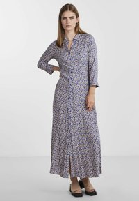 Naine, kes kannab pikka sinist lillelist kleiti, millel on ees nööbid, kolmeveerandikvarrukatega, seisab, ühe käega puusal ja jalas beežid avatud varvastega kingad.