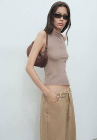 Brown sleeveless top with a high neck, paired with loose beige pants featuring a chain detail. Model holds a small brown bag.