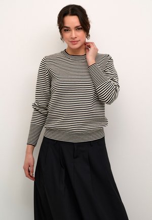 Pull rayé en crème et noir, avec un col rond, des manches longues et un ourlet côtelé. Associé à une jupe plissée noire.