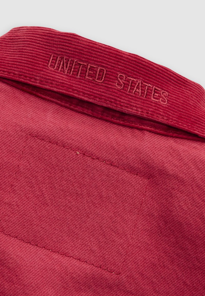 Primo piano di tessuto rosso che mostra un colletto di velluto a coste con la scritta "UNITED STATES" ricamata con filo rosso coordinato.