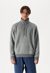 Sudadera gris con cremallera y cuello alto, bolsillo frontal y puños acanalados. Tela suave y texturizada. Parche de logo en la parte inferior delantera.