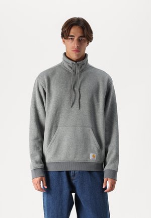 Sudadera gris con cremallera y cuello alto, bolsillo frontal y puños acanalados. Tela suave y texturizada. Parche de logo en la parte inferior delantera.