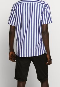 Camicia a maniche corte con strisce verticali blu e bianche, dalla vestibilità comoda e spacchi laterali, abbinata a pantaloncini neri arrotolati.