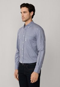 Hombre vestido con una camisa de cuadros azul y blanca de manga larga con botones y jeans oscuros, de pie contra un fondo gris claro liso.