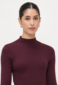 Burgundy ribbien neulepaita korkealla kauluksella ja pitkillä hihoilla, jossa on teksturoitu pinta ja tiukka siluetti.