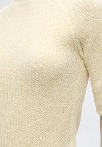 Κοντινό πλάνο ενός χνουδωτού, κρεμ πουλόβερ με κοντά μανίκια, που δείχνει λεπτομερή υφή και ραφές στον ώμο και το στήθος.