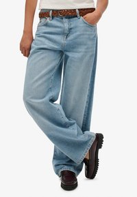 Jasnoniebieskie jeansy o szerokich nogawkach z wyblakłym wykończeniem, wyposażone w pleciony brązowy pasek oraz ciemnofioletowe buty z teksturowaną podeszwą.