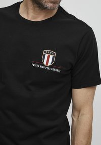 Juodas medvilninis marškinėlis su siuvinėtu šarvo logotipu, kuriame yra metai "1924" ir vertikalios raudonos, baltos bei mėlynos juostelės. Po juo užrašas "FQ1924 HIGH PERFORMANCE."