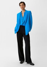 Hellblaues Ein-Knopf-Blazer über einem passenden Oberteil, kombiniert mit schwarzen weit geschnittenen Hosen. Schwarze Schuhe runden das Outfit ab.