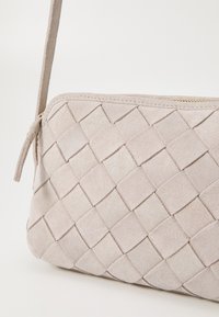 Woven suede torebka kopertówka w jasnym beżu z gładką teksturą, z zamknięciem na suwak i wąskim paskiem na ramię.