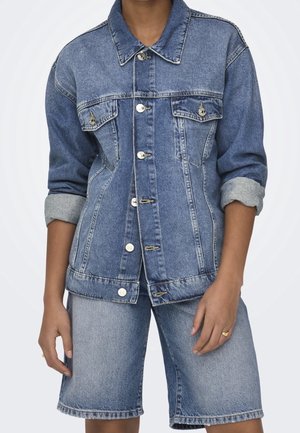Personne portant une veste en denim bleu avec les manches retroussées et un short en denim assorti arrivant aux genoux, sur un fond clair uni.