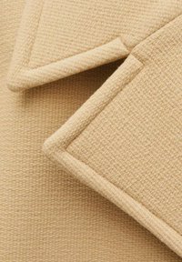 Material textil bej cu un model țesut; prezintă rever ascuțite, în unghi, cu margini curate și detalii subtile de sutură.