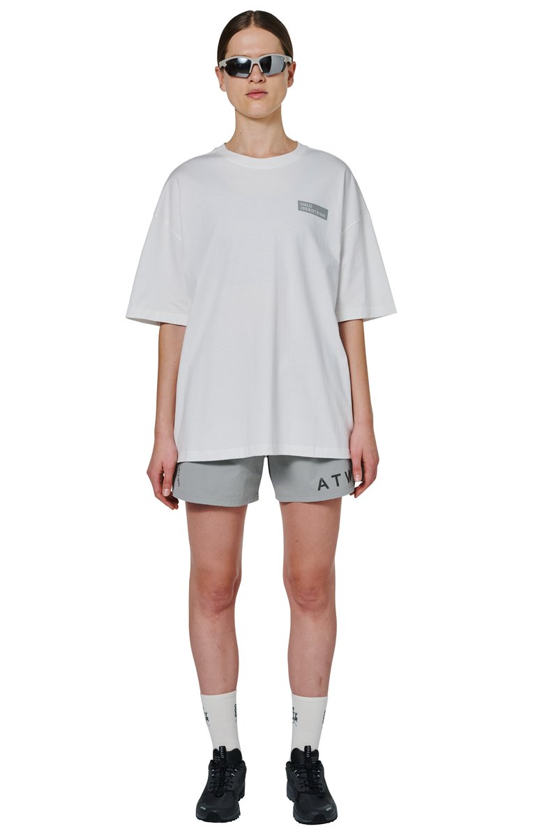 Frau trägt ein übergroßes weißes T-Shirt mit kleinem Brustlogo, hellgraue Shorts mit schwarzer Schrift, weiße Socken, schwarze Sneaker und reflektierende Sonnenbrille.