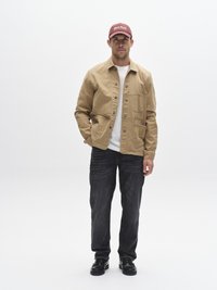 Svetlo rjava platnena jakna z žepi, nošena čez belo majico, kombinirana s temnimi kavbojkami in črnimi čevlji, dopolnjena z bordo kapo.