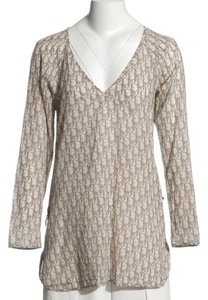Beige bluse med lange ærmer med et lysebrunt geometrisk mønster over det hele og dyb V-hals på en hvid mannequinbuste.