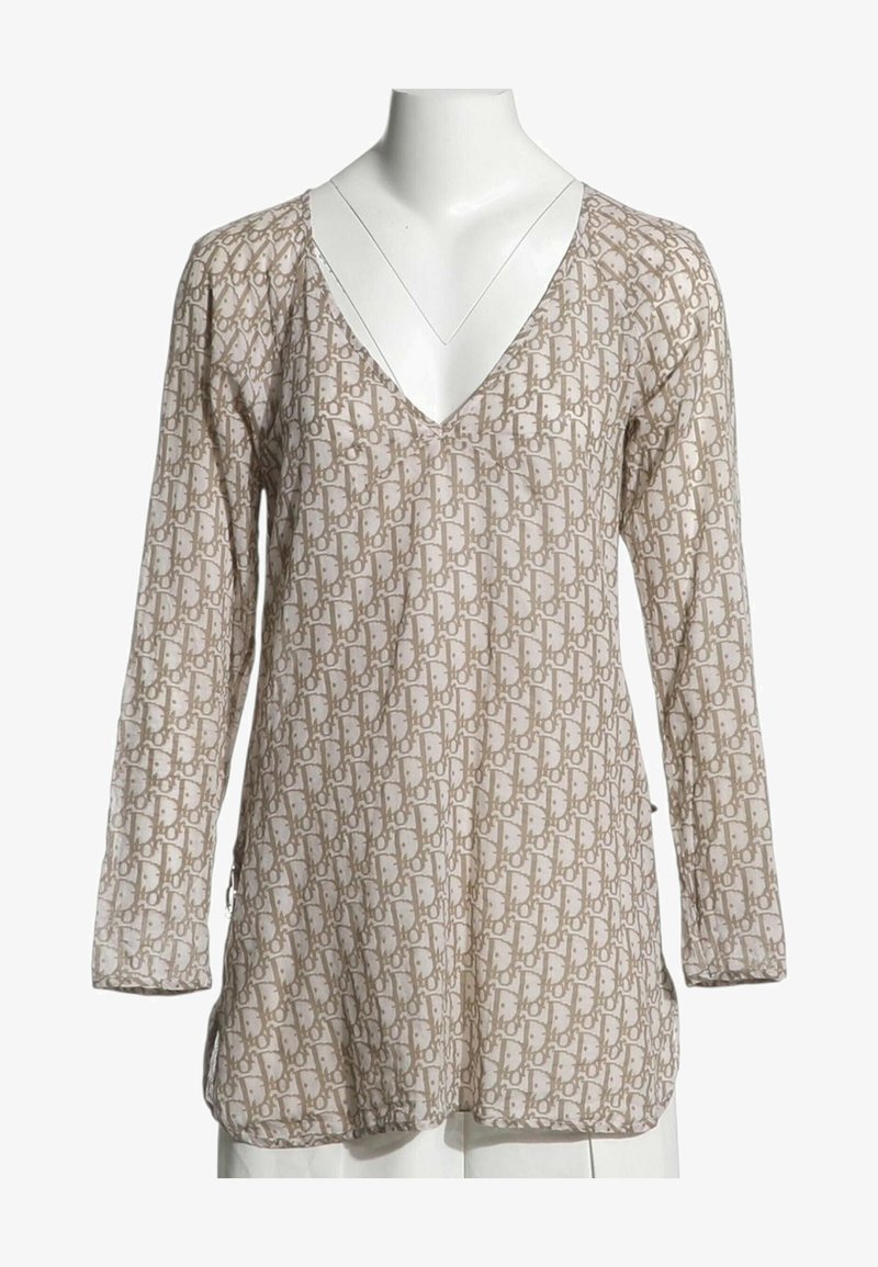 Blouse beige à manches longues avec motif géométrique marron clair sur toute la surface et décolleté en V profond sur un buste de mannequin blanc.