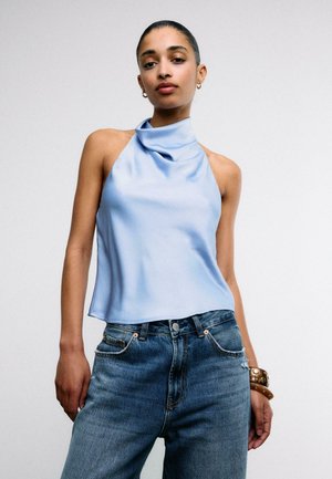 Donna che indossa un top azzurro senza maniche con collo allacciato, jeans blu a vita alta e un braccialetto marrone con borchie al polso.