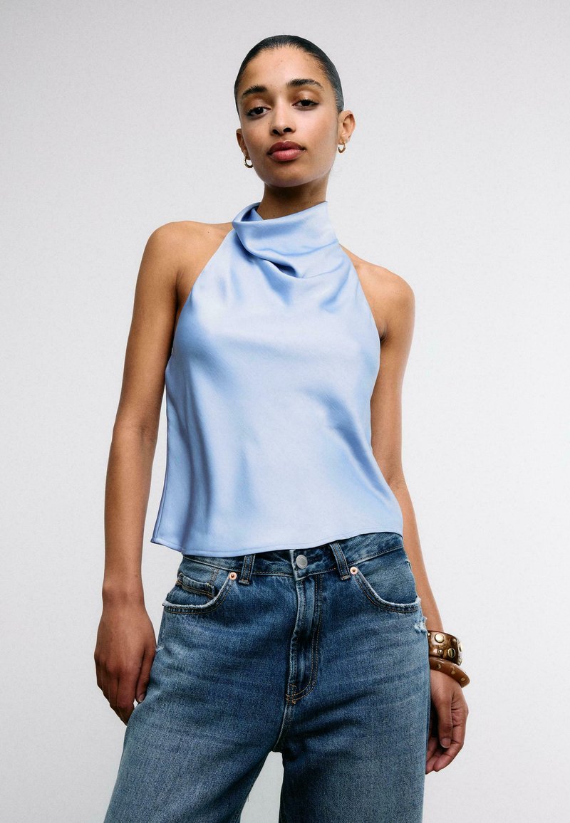 Donna che indossa un top azzurro senza maniche con collo allacciato, jeans blu a vita alta e un braccialetto marrone con borchie al polso.