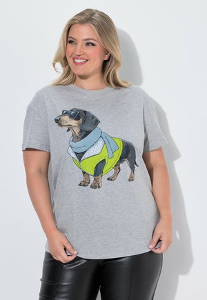 Lächelnde Frau trägt ein graues T-Shirt mit einer Grafik eines Dackels mit Sonnenbrille, Schal und gelber Jacke.