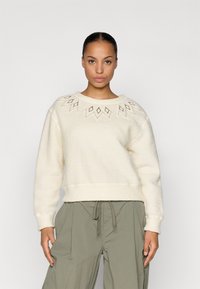 Sudadera de color crema con cuello redondo, que presenta patrones de diamantes bordados a lo largo del cuello. Fabricada con un tejido suave y texturizado.