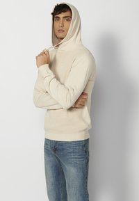 Sweat à capuche beige avec un motif de tricot texturé, cordon de serrage au cou, poignets et ceinture côtelés. Associé à un jean bleu clair.