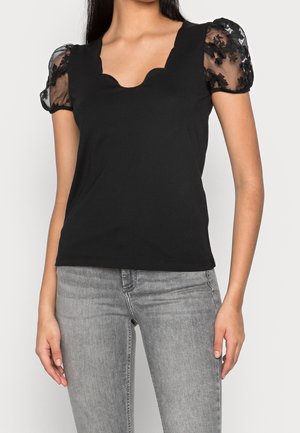 Kobieta w czarnej bluzce z falbaniastym dekoltem i przejrzystymi bufkami z haftem kwiatowym, w zestawieniu z jasnoszarymi dżinsami.