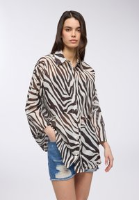 Camicia oversize a stampa zebra in tessuto trasparente con strisce marroni e crema, caratterizzata da un colletto classico e chiusura con bottoni davanti, abbinata a pantaloni corti di jeans.