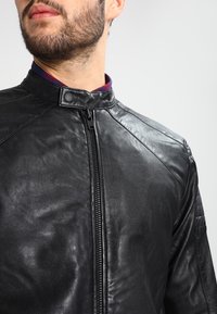 Black leather jacket with a round collar, zip front, and quilted shoulder details. Smooth texture and fitted design enhance its structure.