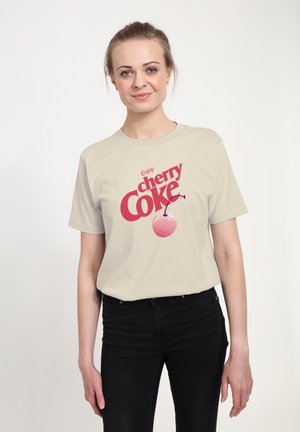 Světle béžové tričko s červeným grafickým textem "Užijte si cherry Coke" a proužkovaným designem třešně. Nosené s černými džíny, na jednobarevném pozadí.