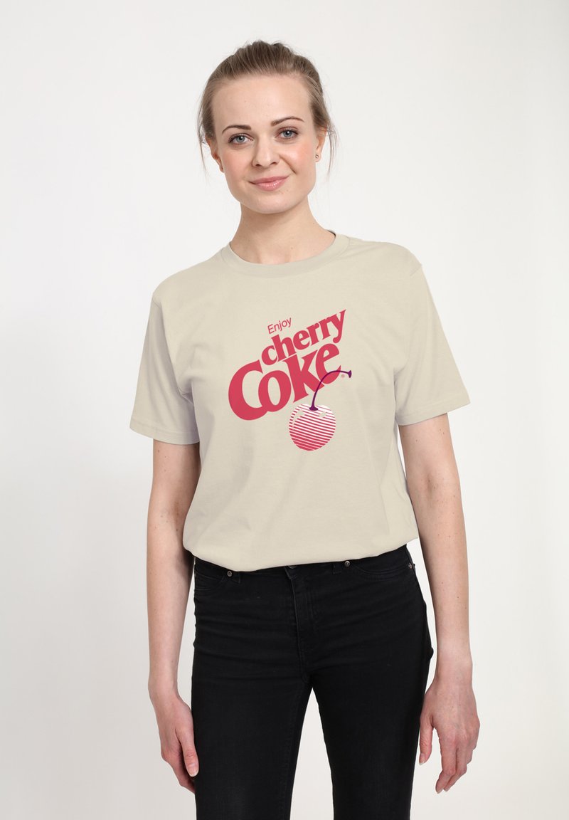 Světle béžové tričko s červeným grafickým textem "Užijte si cherry Coke" a proužkovaným designem třešně. Nosené s černými džíny, na jednobarevném pozadí.