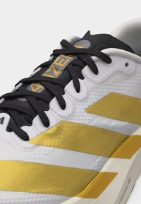 Zapatilla deportiva blanca y dorada con cordones negros y "AZE" visible en la lengüeta negra en texto amarillo.