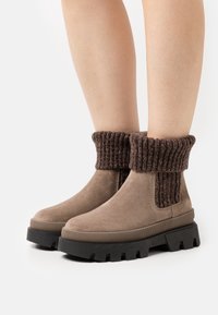 Brązowe zamszowe botki za kostkę z prążkowanym ciemno-brązowym ścianką i czarną grubą podeszwą. Gładka tekstura i prosty design.