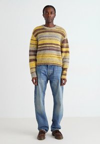Pull en tricot rayé en jaune, marron et gris, avec un col rond. Associé à un jean en denim bleu clair et des chaussures marron.